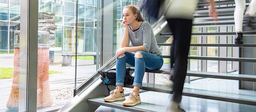 A young women sits on the steps in her college looking worried whilst other students walk past her