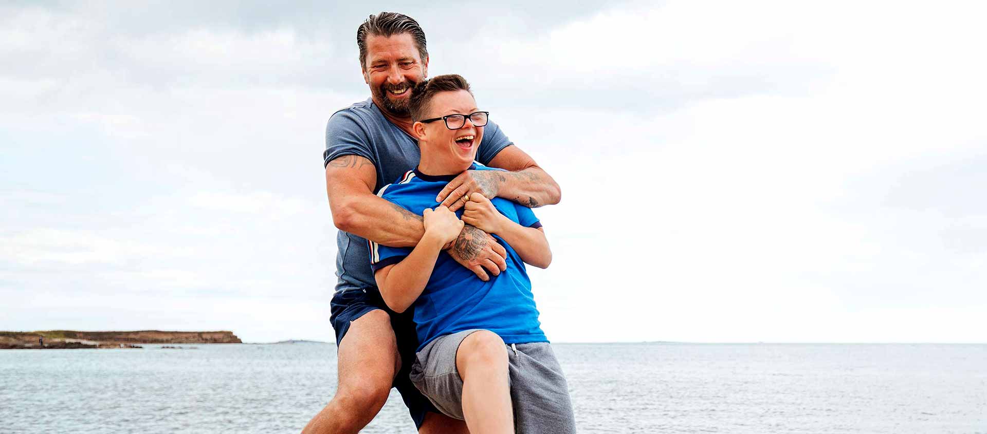 A happy father cuddles his young son at the beach. There are cliffs and the sea in the background.