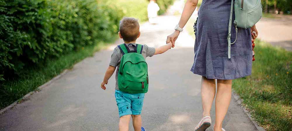 A young boy with a green backpack is holding his mother's hand as they are walking through a park.