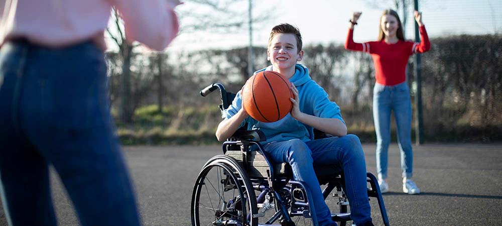 A young boy is sitting in his wheel chair holding a basketball. There is an adult and a young girl around him. They are all on an outdoor basketball court.