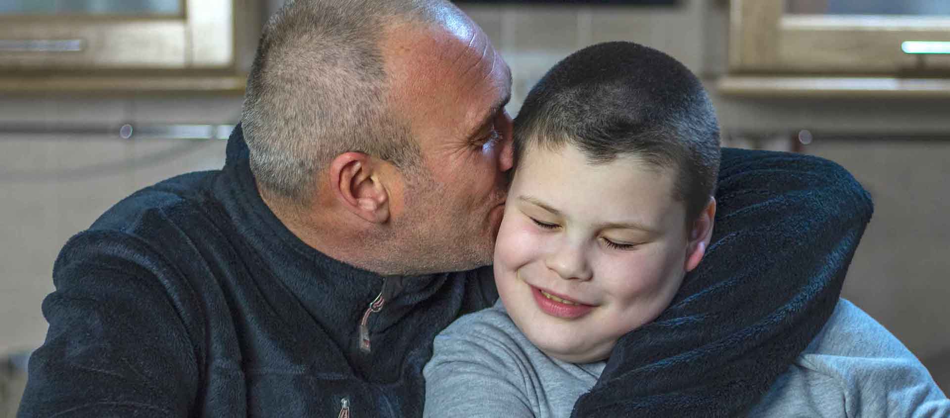 A father cuddles his son and is kissing him on the cheek.