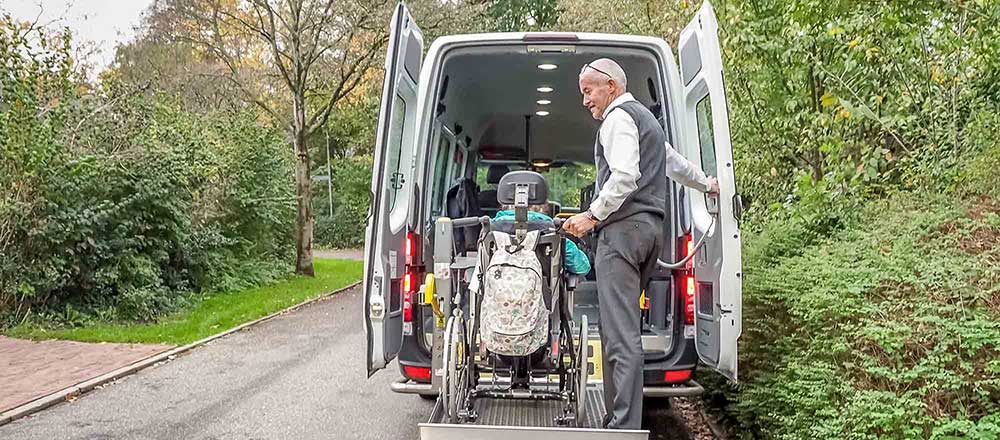A man helps a young child in a wheel chair into the back of the school minibus