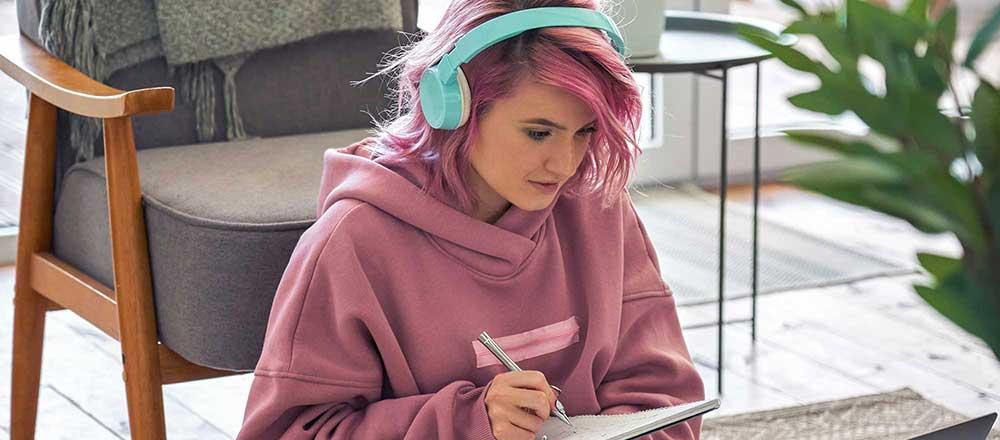 A young lady with a pink hoodie and pale blue headphones is working at a laptop.