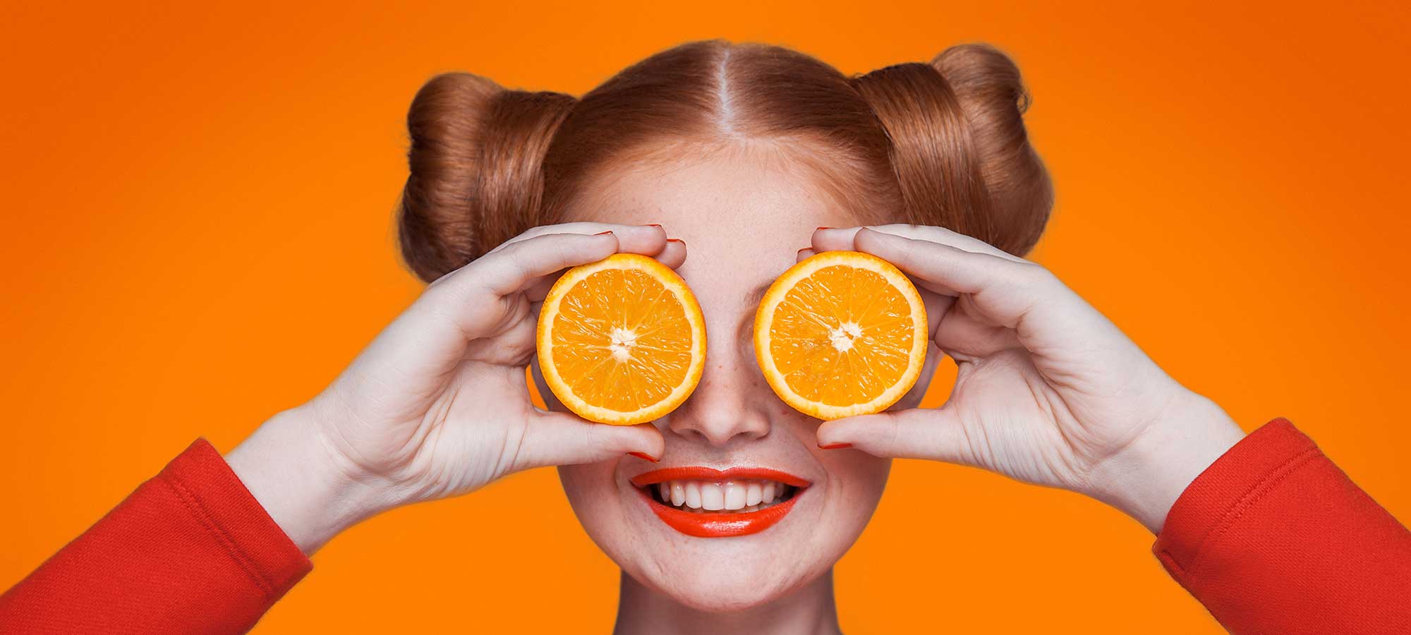 A lady has two halves of an orange held in front of her eyes. She has reddish hair and is smiling. There is a bright orange background.