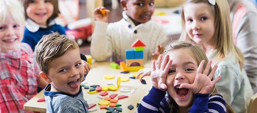 Young children are sitting around a school desk. Two children in the front are making silly faces.