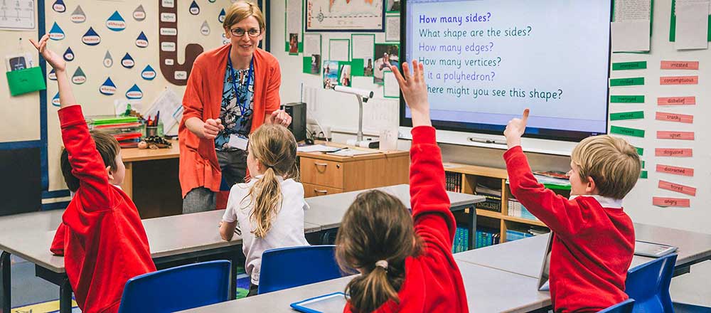 A group of school children are sitting on the floor of their classroom. The teacher is sat on her desk, asking them a question