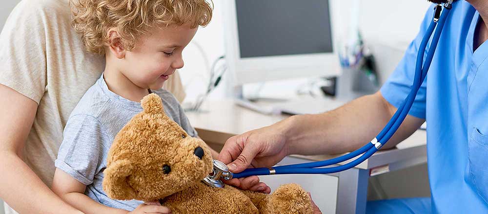 A doctor puts his stethoscope onto a young boys teddy bear. The child is holding the bear and smiling.