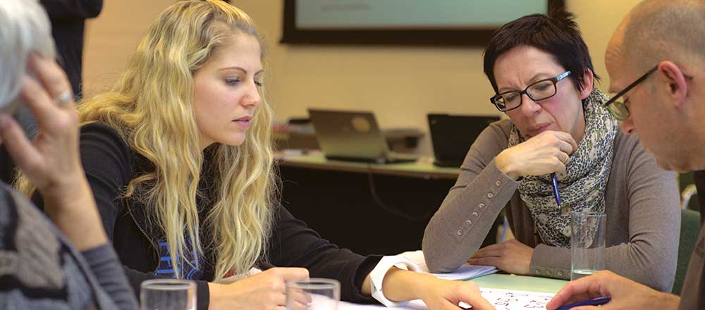 A group of professional women gather around a table. Each woman has a laptop.