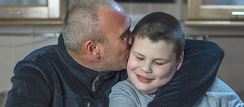 A father cuddles his son and is kissing him on the cheek.