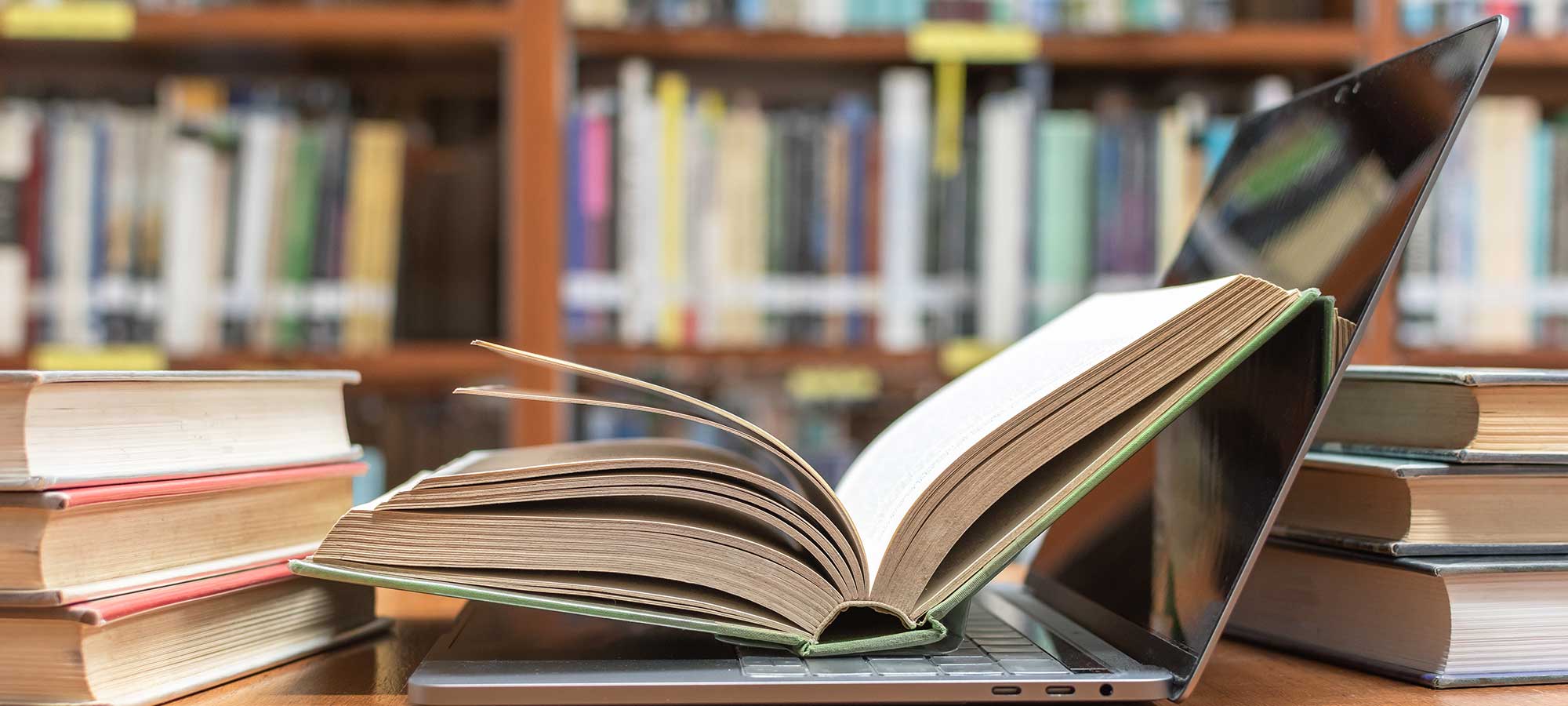 A pile of text books surrounds a laptop PC. There is an open book lying on the laptop. There are rows of books in the background.