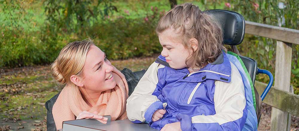 A woman is crouched by the wheel chair of a disabled girl. They are smiling at each other