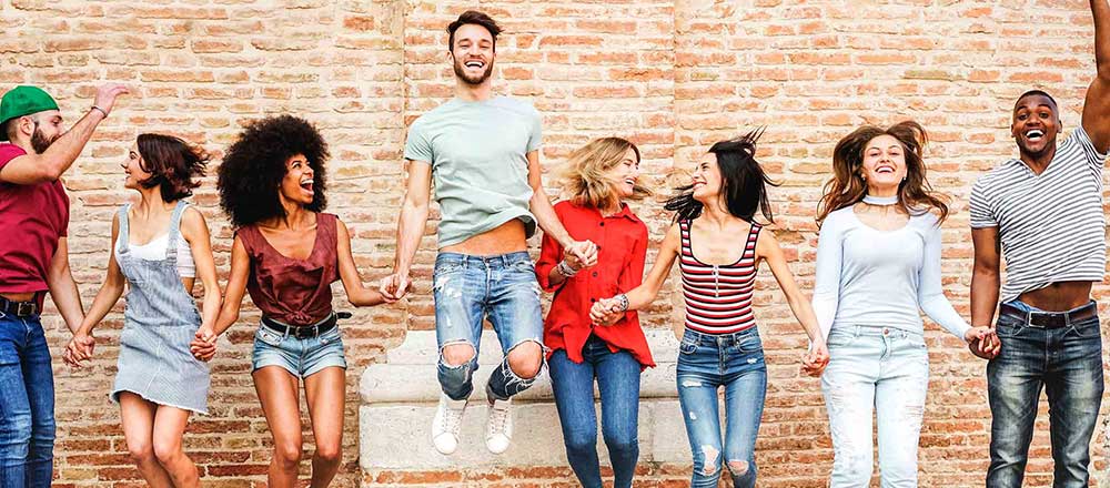 A group of young people are all jumping in the air and laughing. There is a brick wall behind them.