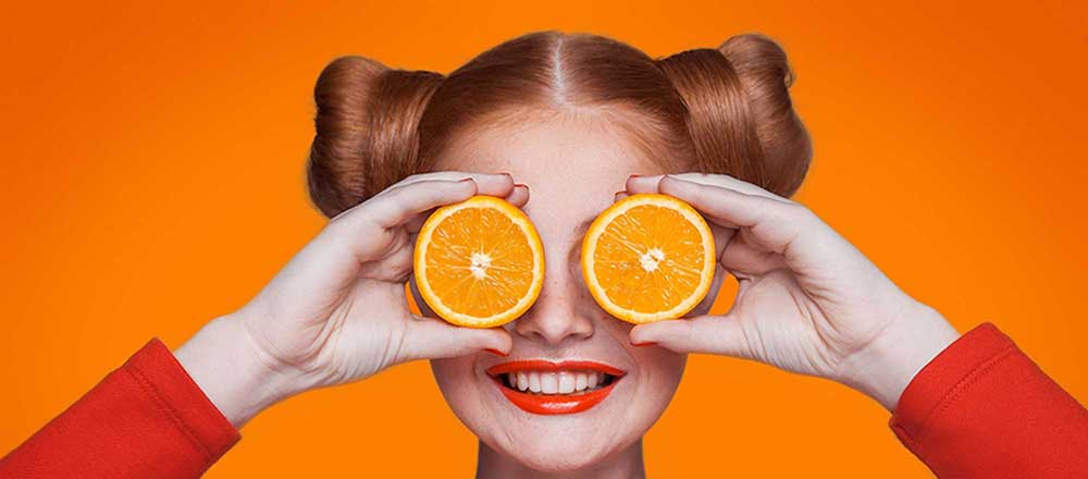 A lady has two halves of an orange held in front of her eyes. She has reddish hair and is smiling. There is a bright orange background.